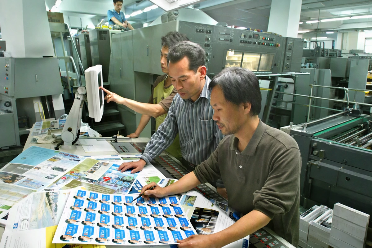 Workers inspect freshly printed label sheets on a table inside an offset printing workshop, checking uv varnish coverage and registration quality next to a large printing press Workers inspect freshly printed label sheets on a table inside an offset printing workshop, checking uv varnish coverage and registration quality next to a large printing press